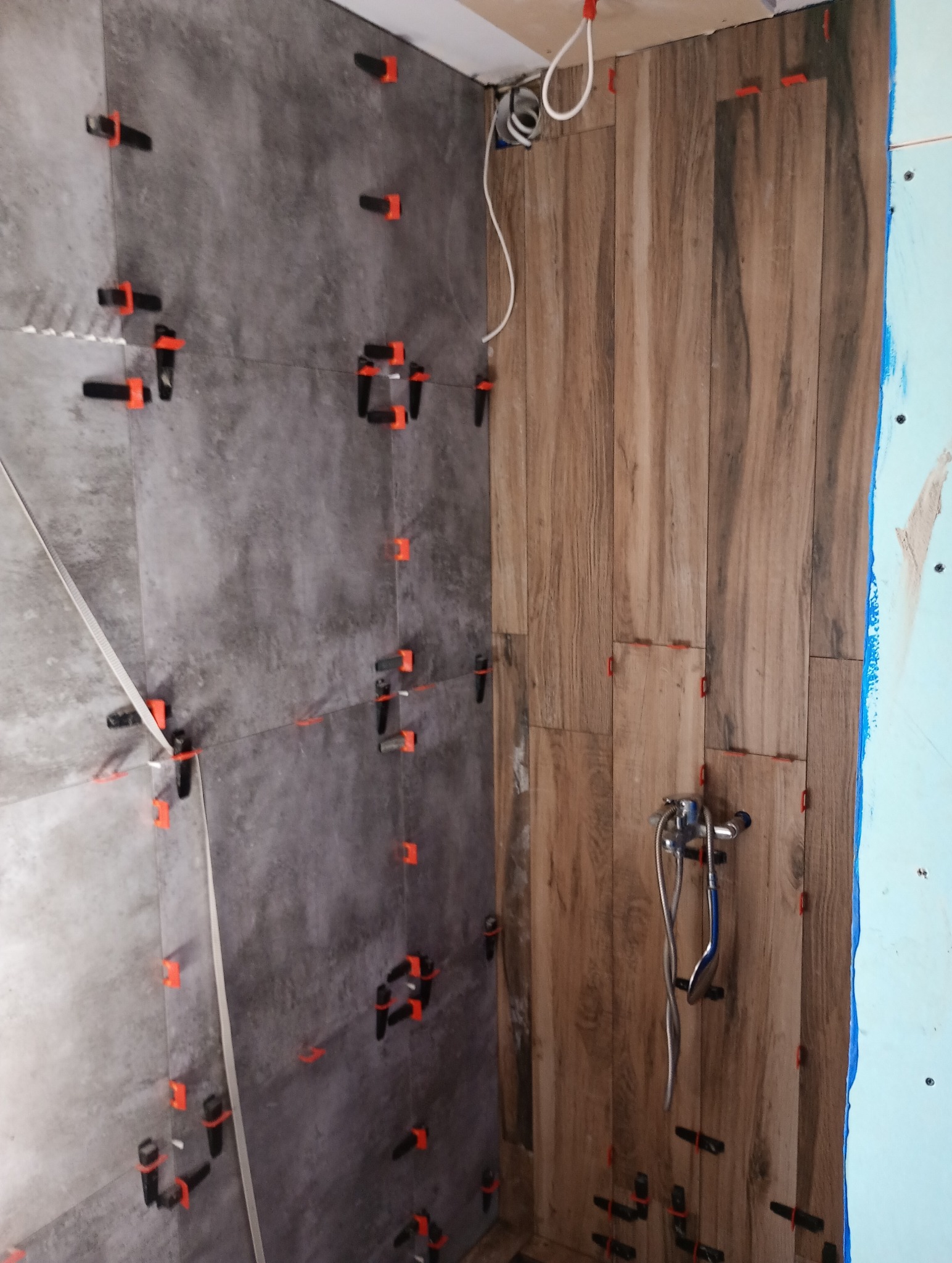 Montaż płytek imitujących drewno i szarych płytek na ścianach prysznica, z widocznymi krzyżykami dystansowymi i poziomnicą.