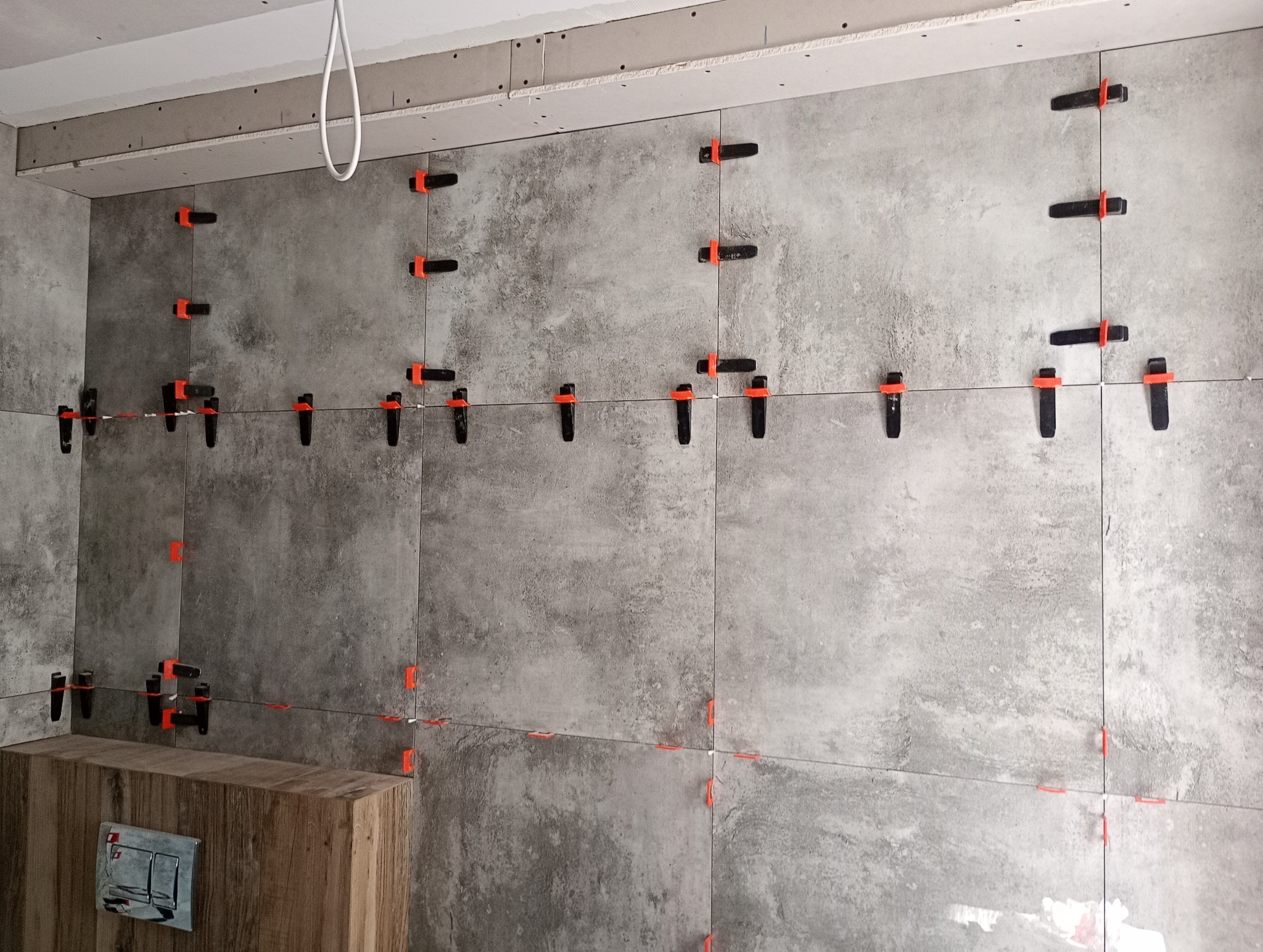 Ściana w trakcie układania szarych płytek imitujących beton, z użyciem czarnych klipsów i pomarańczowych klinów poziomujących, widoczny fragment zabudowy stelaża WC.