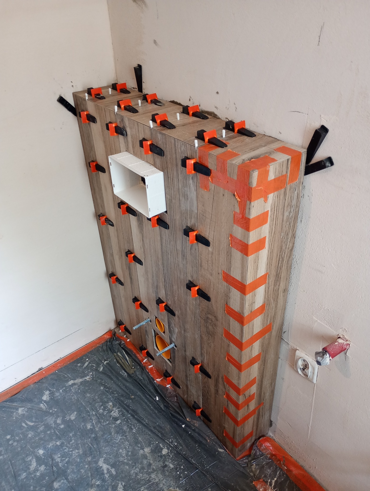 Montaż płytek imitujących drewno na narożniku ściany w łazience, z użyciem klipsów poziomujących i taśmy malarskiej, widoczne puszki instalacyjne i zabezpieczona folią podłoga.