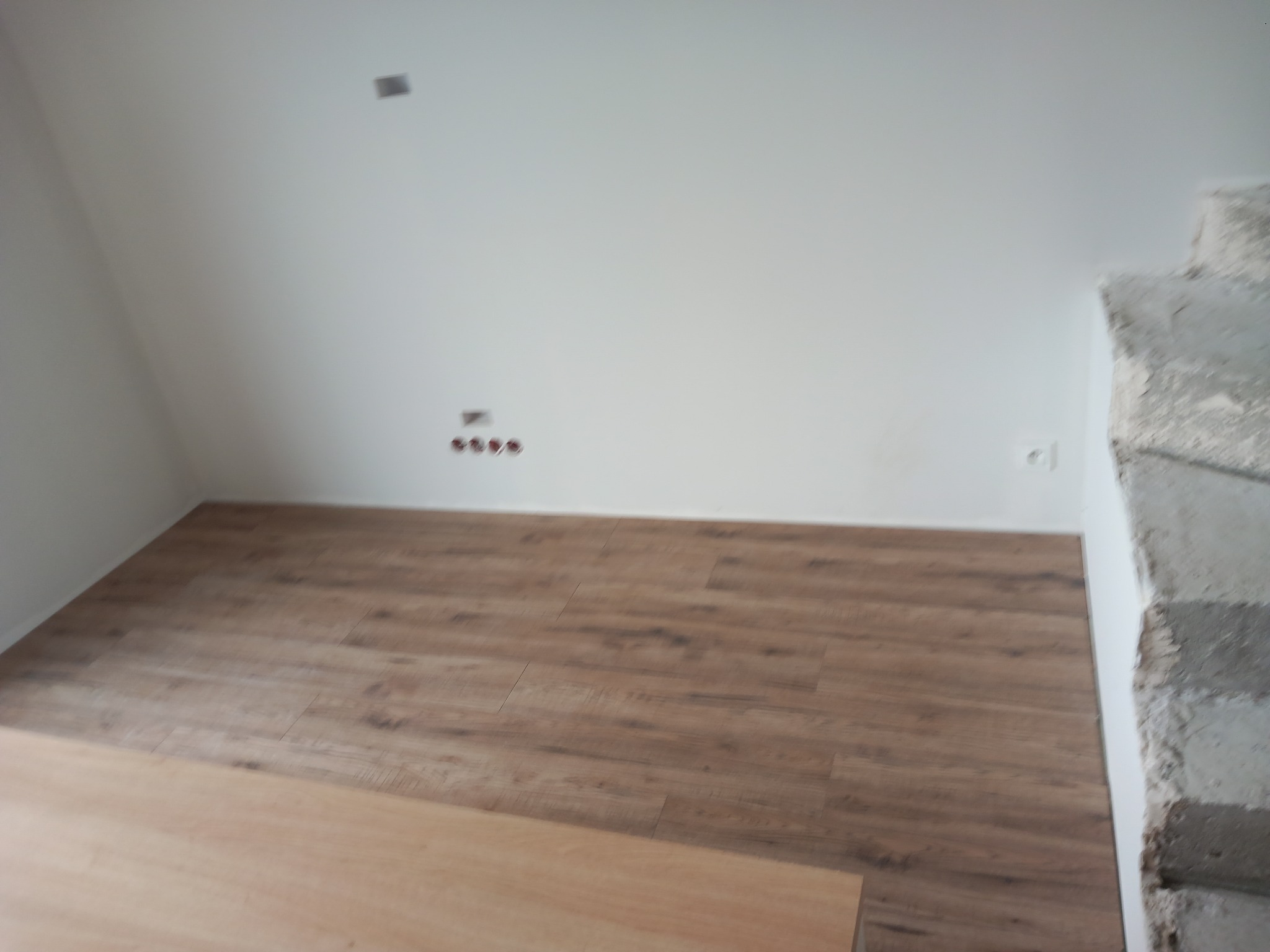 Fragment pokoju z panelami podłogowymi w odcieniach brązu, białe ściany z widocznymi gniazdkami elektrycznymi i puszkami instalacyjnymi, oraz surowy betonowy element konstrukcyjny przy ścianie.