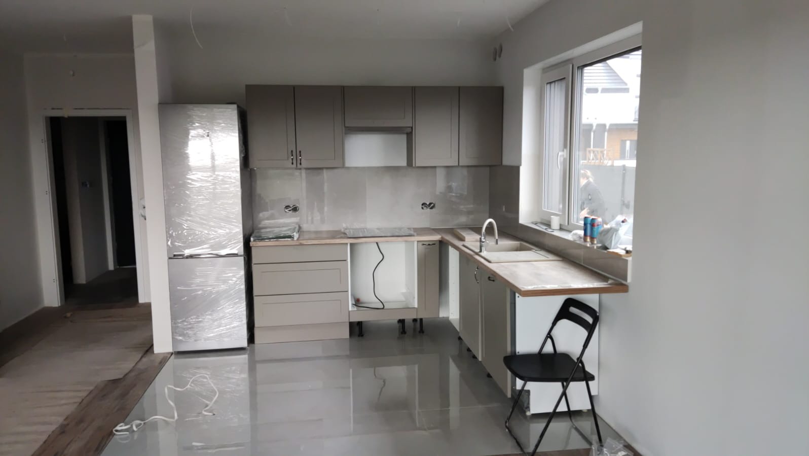 Wnętrze kuchni w trakcie montażu: szare szafki kuchenne, blat z imitacji drewna, zlewozmywak, lodówka owinięta folią ochronną, płytki podłogowe z połyskiem, widoczne kable i puszki elektryczne.