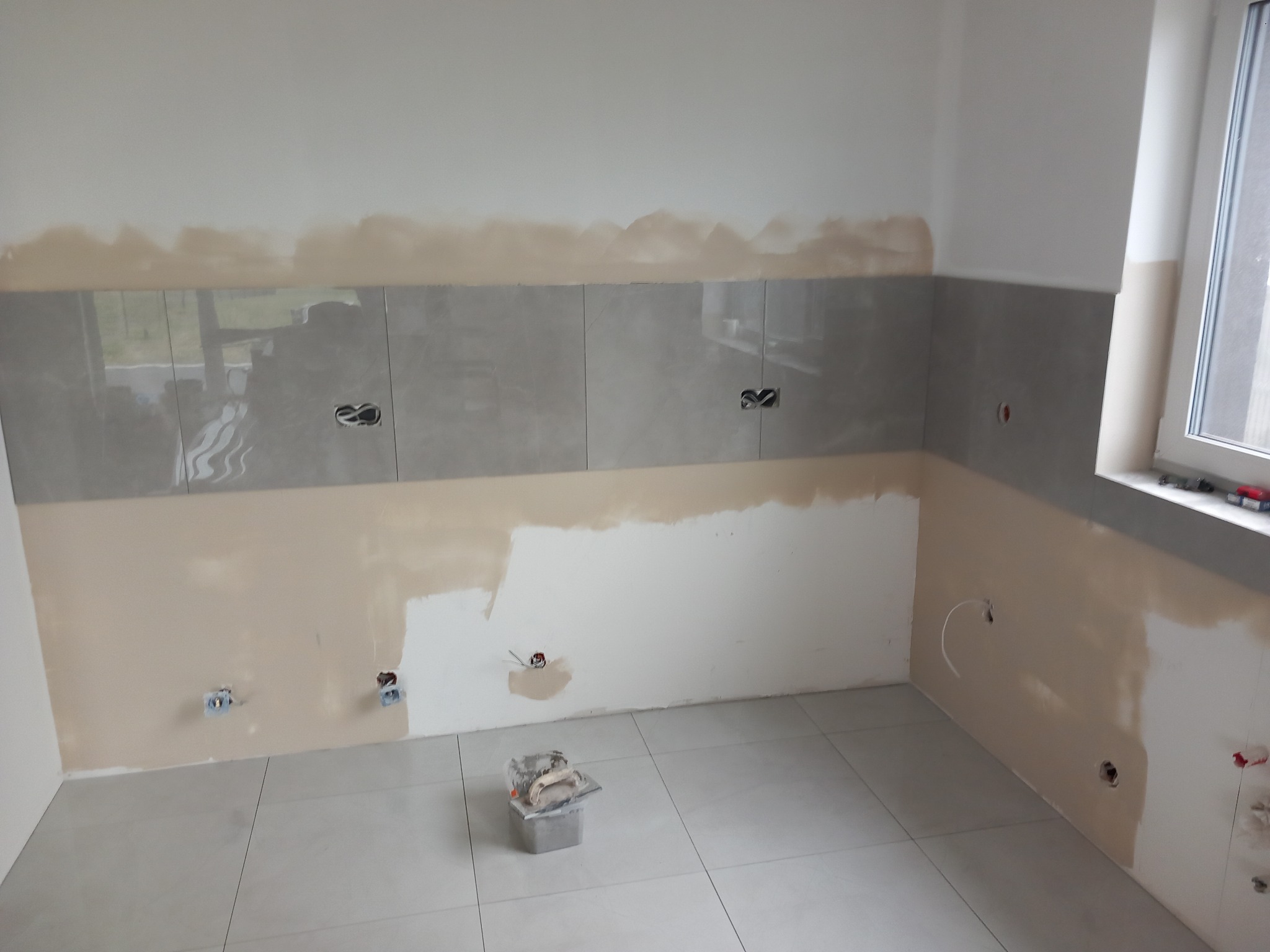 Wnętrze łazienki w trakcie prac: częściowo położone szare płytki na ścianie, ślady po położeniu warstwy wyrównującej podłoże, widoczne puszki elektryczne, na podłodze wiadro z narzędziami.