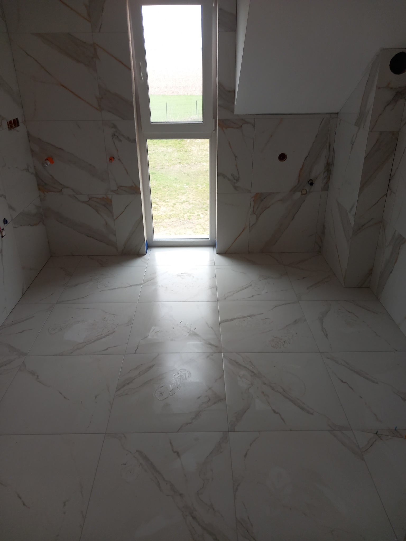 Wnętrze łazienki z białymi płytkami imitującymi marmur na ścianach i podłodze, okno w głębi pomieszczenia, widoczne otwory na instalacje elektryczne i wodne.