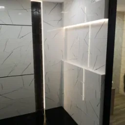 Ściana wyłożona białymi płytkami imitującymi marmur z czarnymi żyłkami, oświetlona listwą LED. Na ścianie wstawka z ciemnych płytek w kształcie heksagon&oacute;w. Podłoga wyłożona czarnymi płytkami.