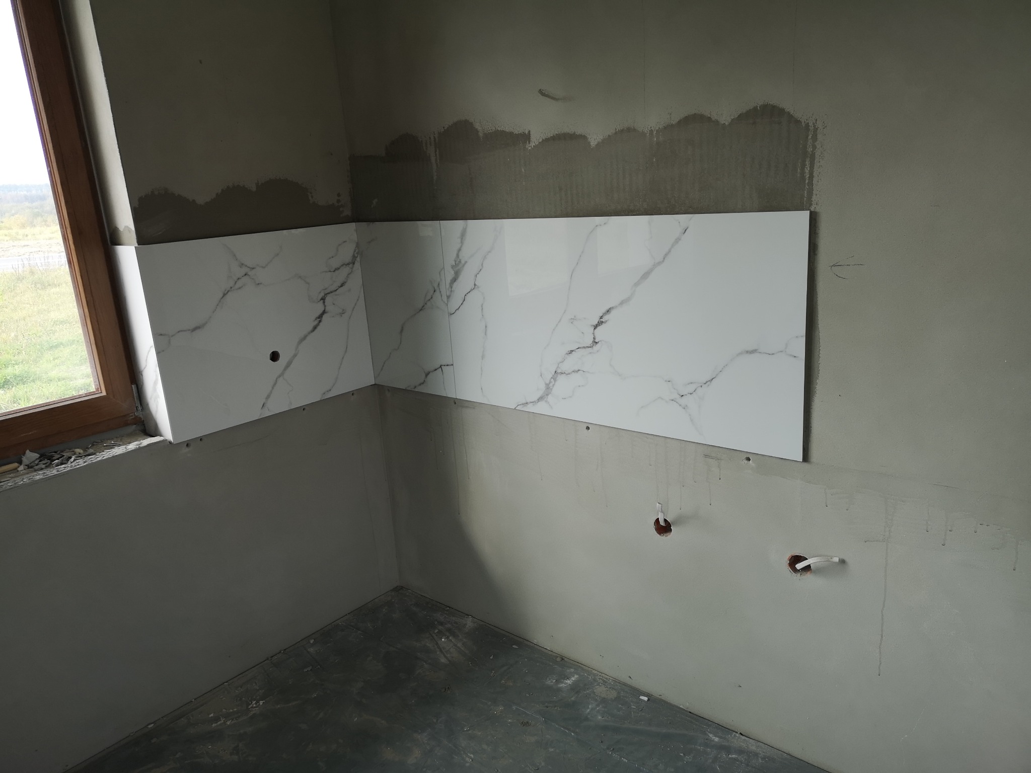 Fragment ściany z częściowo ułożonymi białymi płytkami imitującymi marmur; widoczne wystające przewody instalacji elektrycznej i niezakończone prace wykończeniowe.