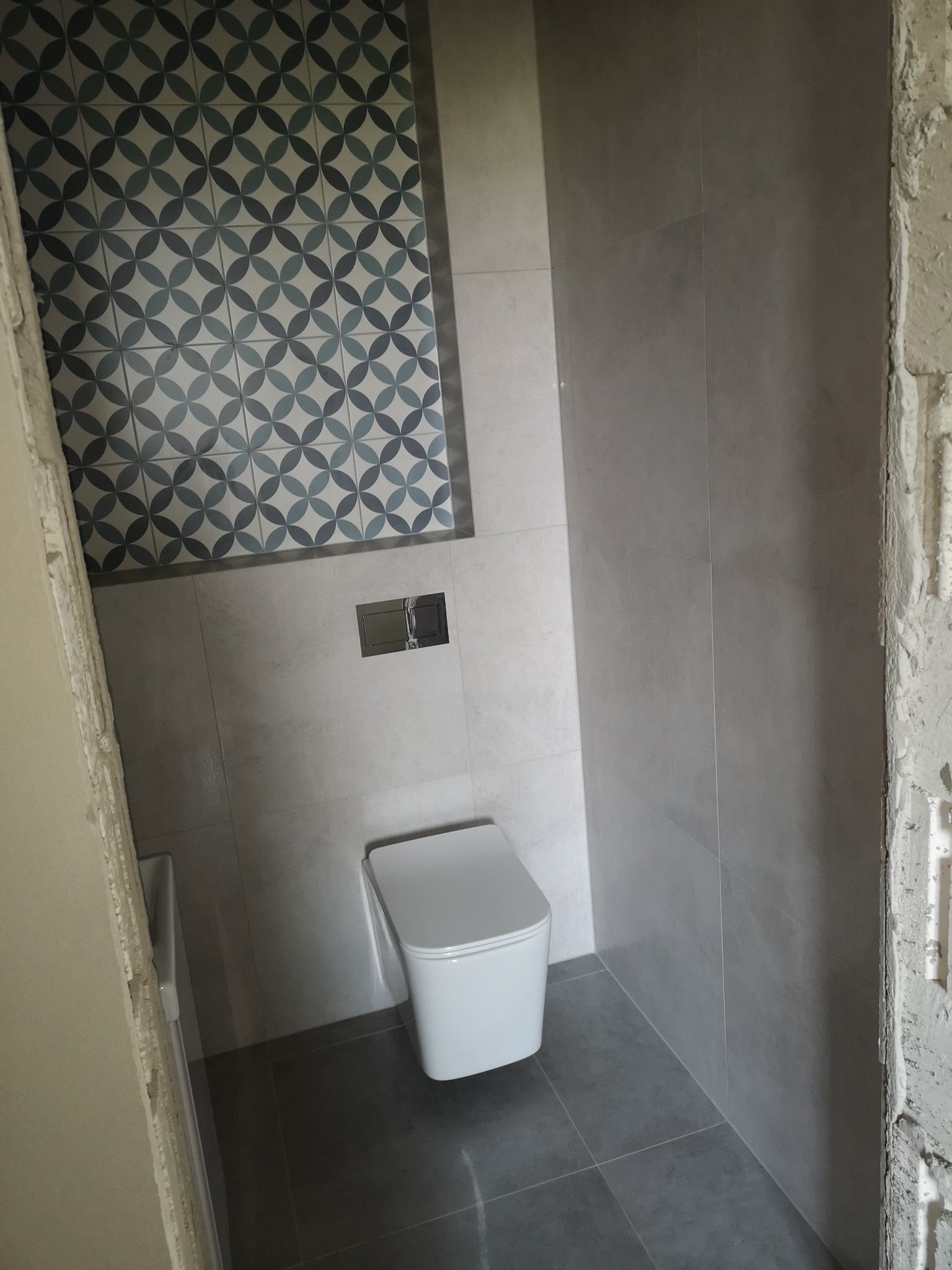 Wnętrze łazienki w trakcie remontu: szare płytki na ścianach i podłodze, dekoracyjne płytki z motywem kwiatowym nad toaletą, widoczne surowe wykończenie ściany.