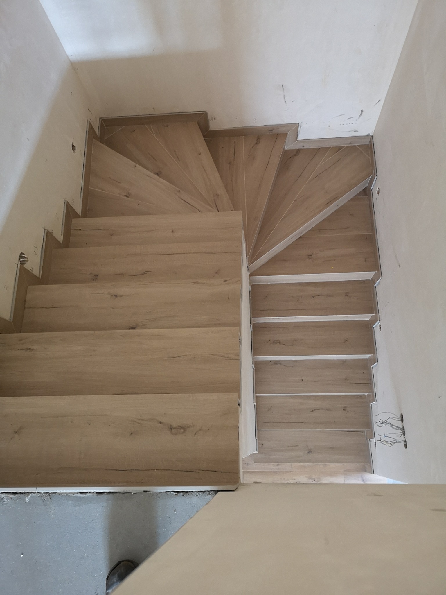 Widok z góry na drewniane schody z panelami, z widocznymi aluminiowymi profilami krawędziowymi, ściany w stanie surowym z wystającymi kablami elektrycznymi, ukazujące etap wykończenia wnętrza.