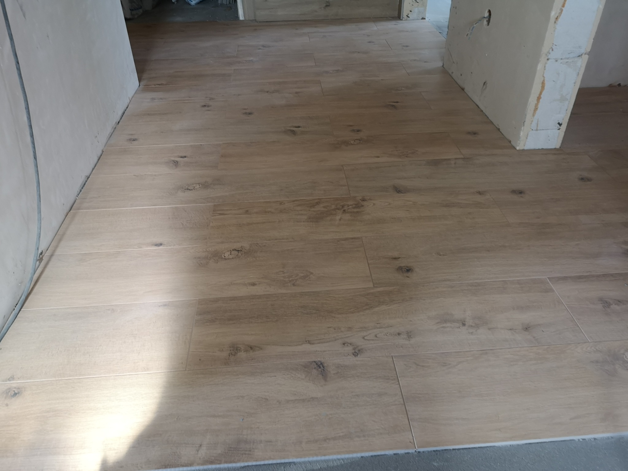 Beżowe płytki podłogowe imitujące drewno, ułożone w nowym pomieszczeniu z surowymi ścianami i widocznymi kablami.