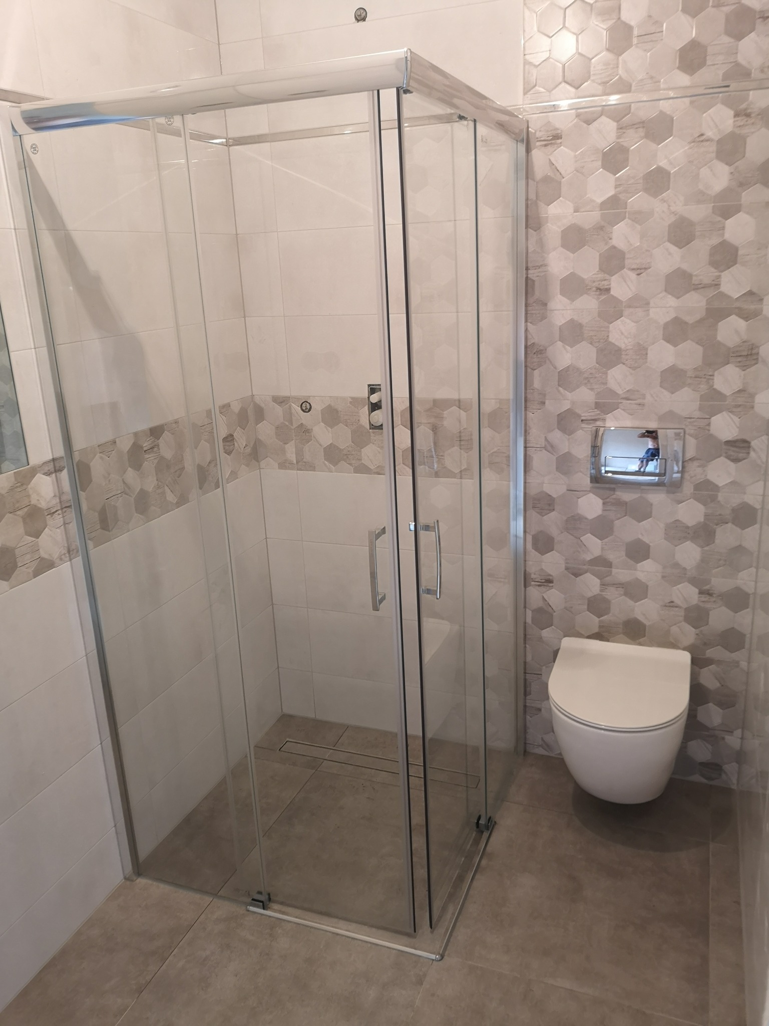 Nowoczesna kabina prysznicowa z odpływem liniowym, jasne płytki heksagonalne na ścianie, podwieszana toaleta.