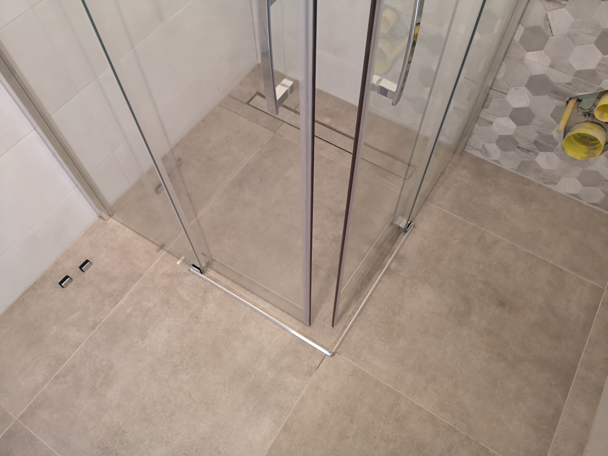 Szklana kabina prysznicowa narożna z chromowanymi uchwytami, beżowe płytki podłogowe i fragment ściany z szarymi heksagonami w tle.