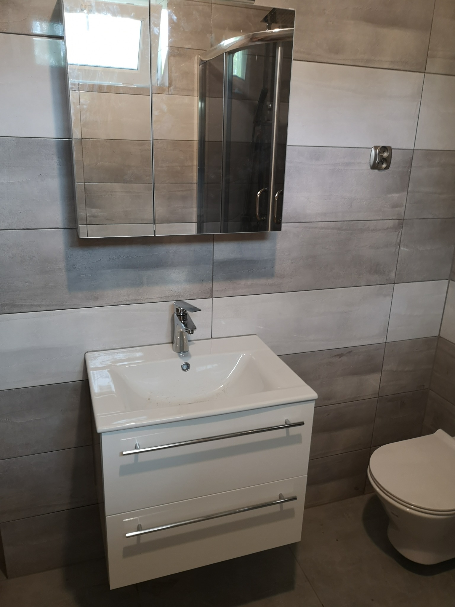 Umywalka z szafką i lustrem w nowocześnie wykończonej łazience z szarymi, poziomymi płytkami imitującymi kamień, widoczna kabina prysznicowa i toaleta.