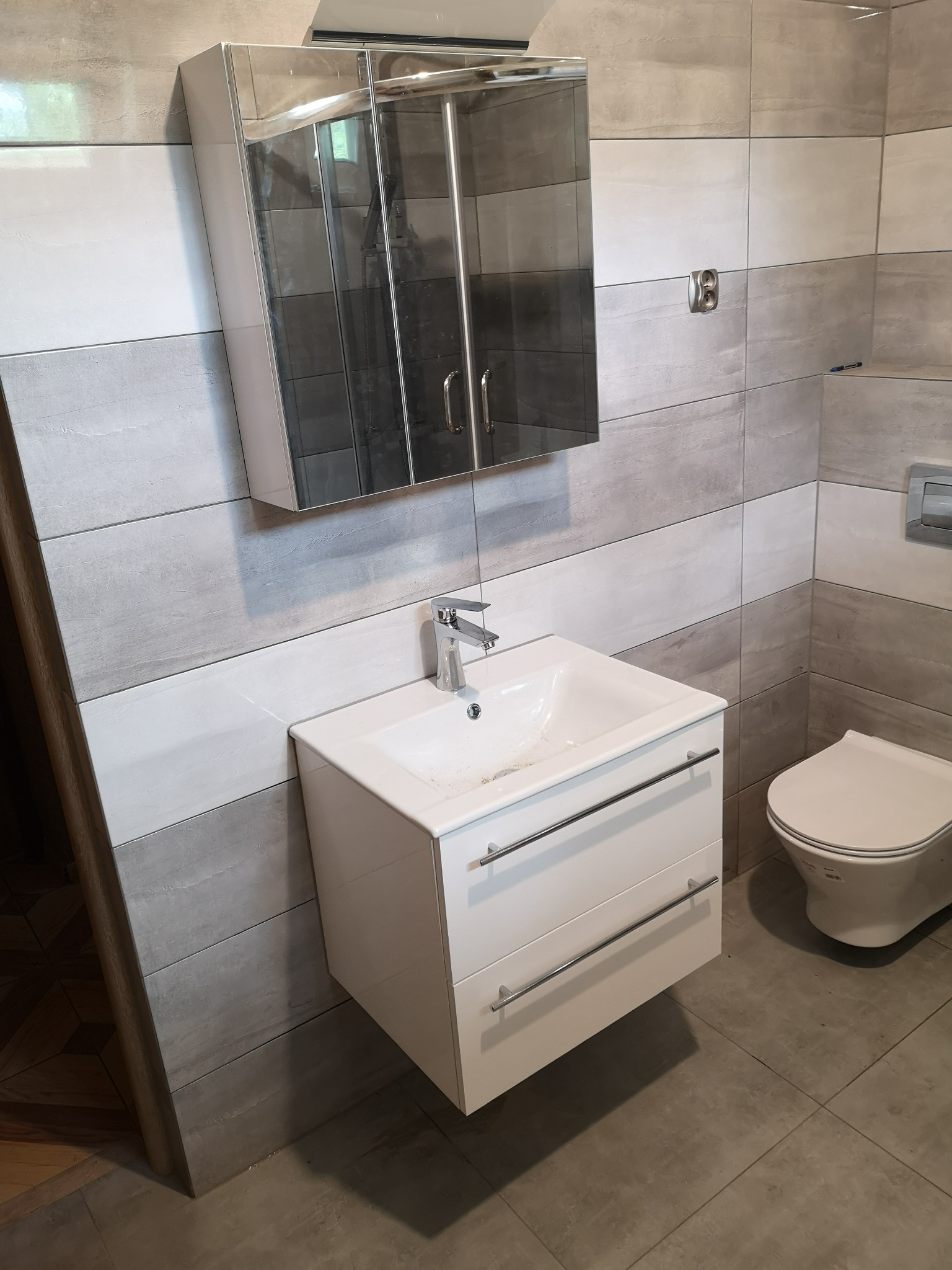 Nowoczesna łazienka z szaro-białymi płytkami imitującymi drewno, białą szafką pod umywalką z chromowanymi uchwytami i lustrzaną szafką wiszącą, widoczny fragment toalety.