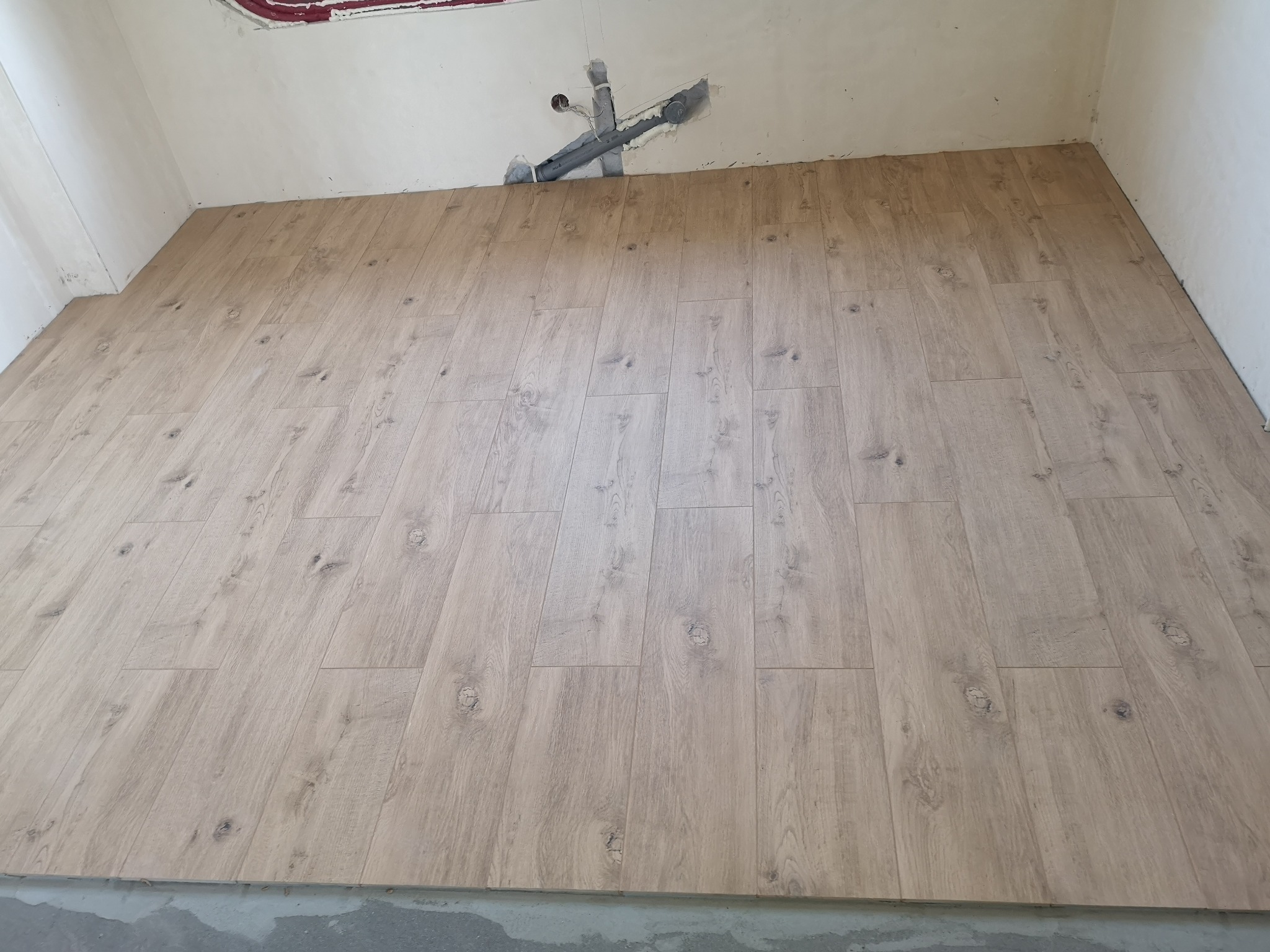 Ułożone panele podłogowe w jasnym odcieniu drewna, widoczne rury wystające ze ściany w górnej części kadru, ściany w trakcie wykończenia.