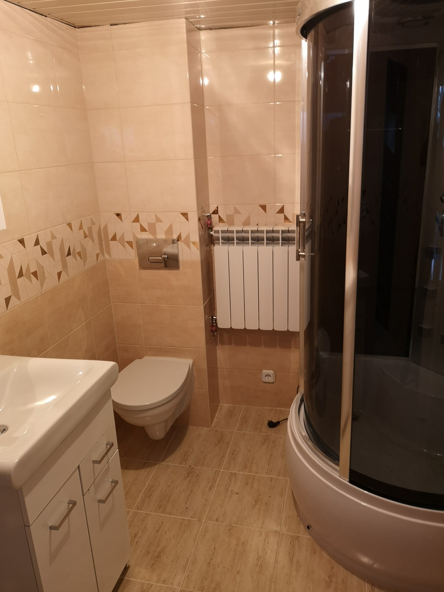 Jasna łazienka z beżowymi płytkami, dekoracyjnym pasem z geometrycznym wzorem, białą umywalką z szafką, toaletą, grzejnikiem i kabiną prysznicową z ciemnymi szybami.