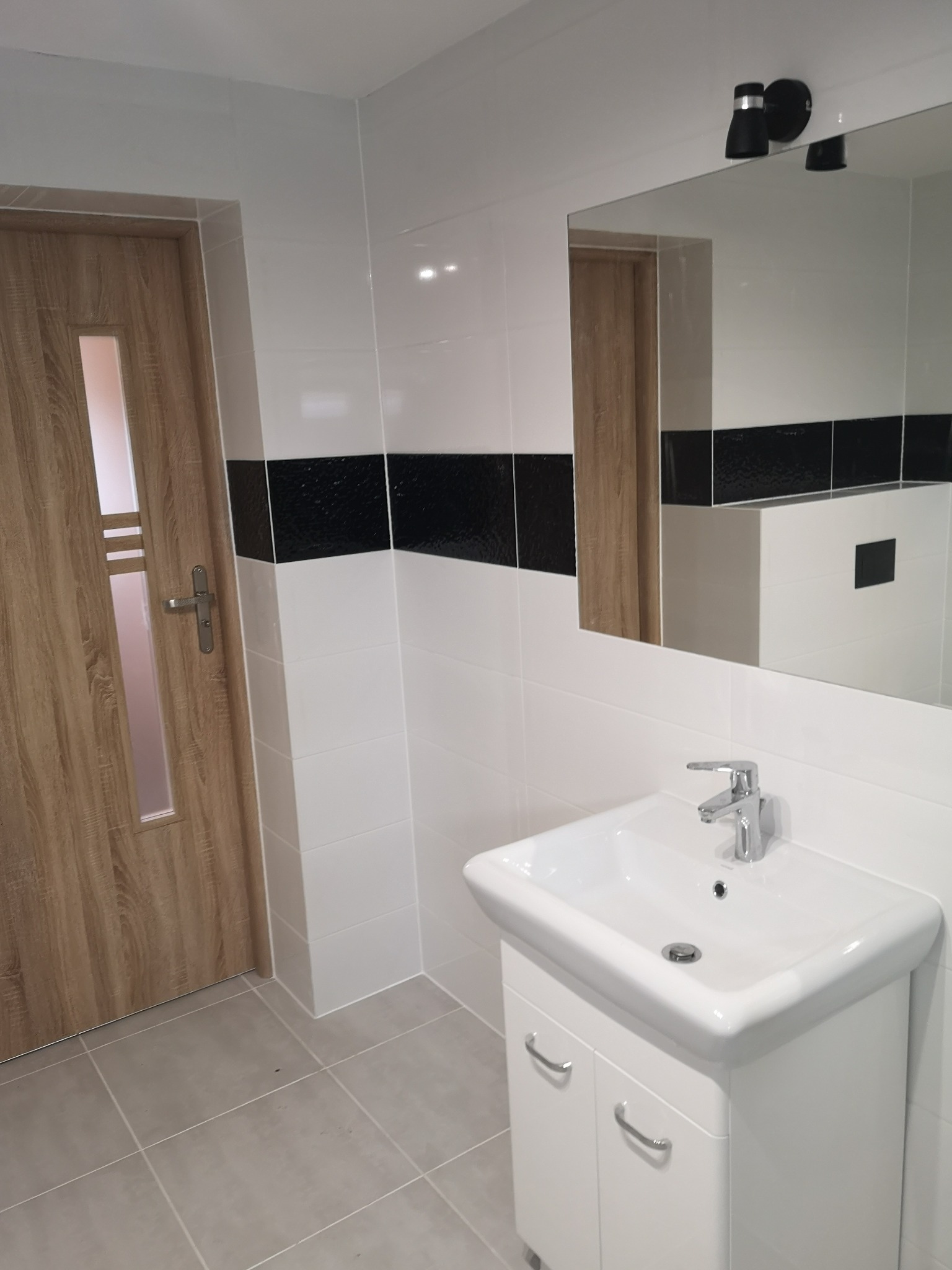 Nowoczesna łazienka po remoncie: białe płytki z czarnym pasem dekoracyjnym, umywalka z szafką, lustro z oświetleniem punktowym i drewniane drzwi z szybką.