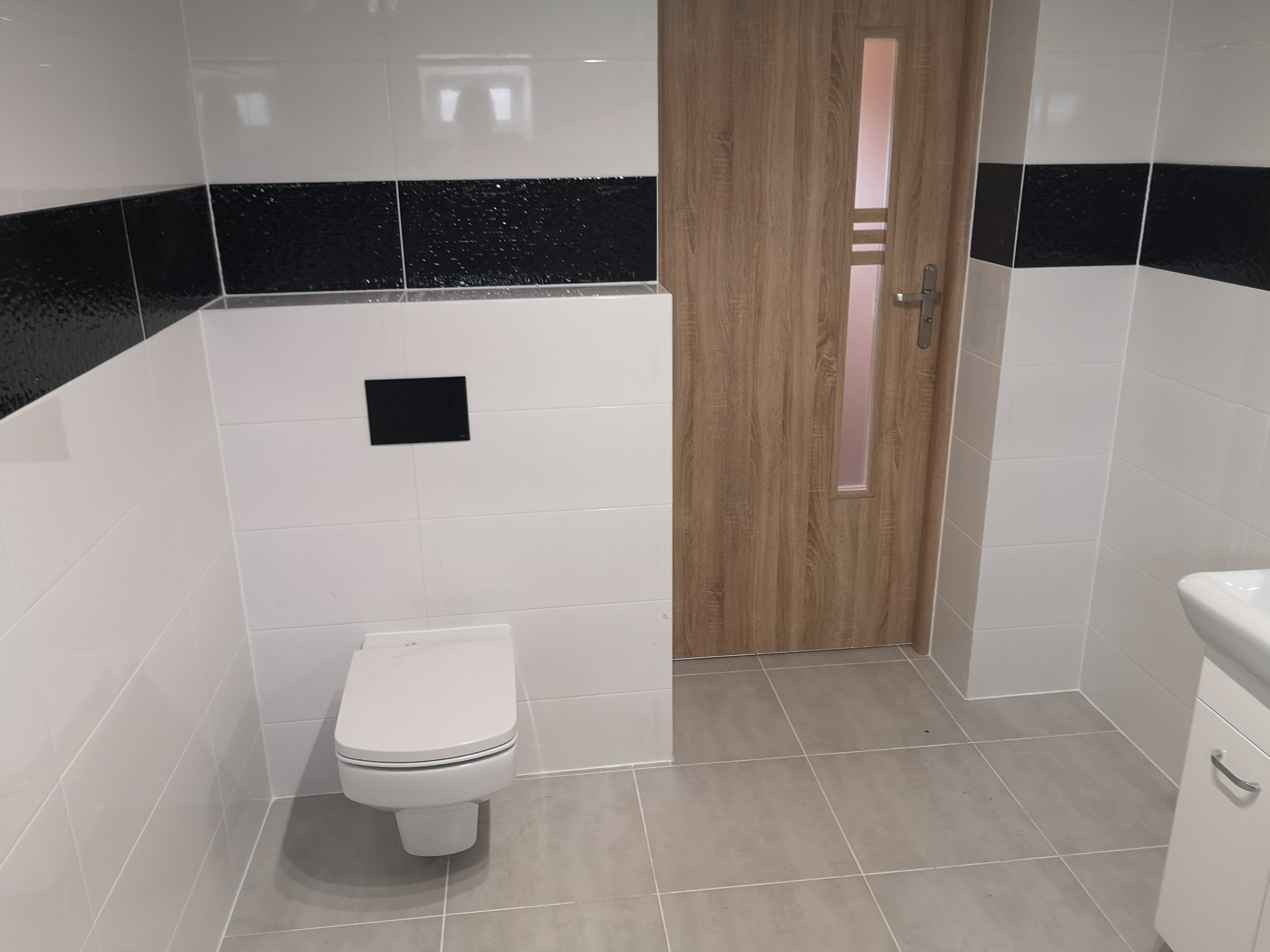 Wykończona łazienka z białymi i czarnymi płytkami na ścianach, wisząca toaleta i drewnopodobne drzwi z szybą.