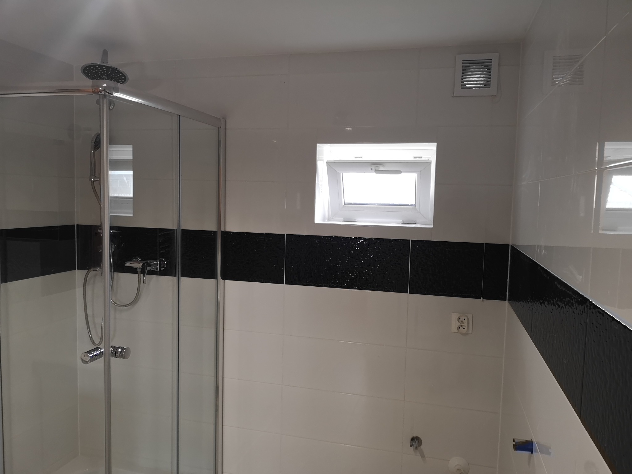 Nowoczesna łazienka z kabiną prysznicową, białymi płytkami i czarnym pasem dekoracyjnym, widoczne okno dachowe i kratka wentylacyjna.