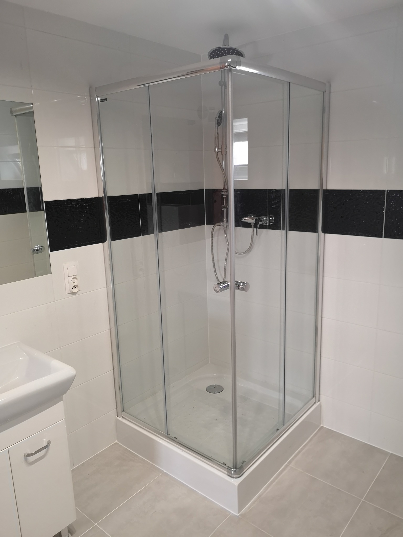 Nowoczesna kabina prysznicowa z chromowanymi profilami i deszczownicą, zamontowana w łazience z białymi płytkami i czarnym pasem dekoracyjnym, widoczna umywalka z szafką.