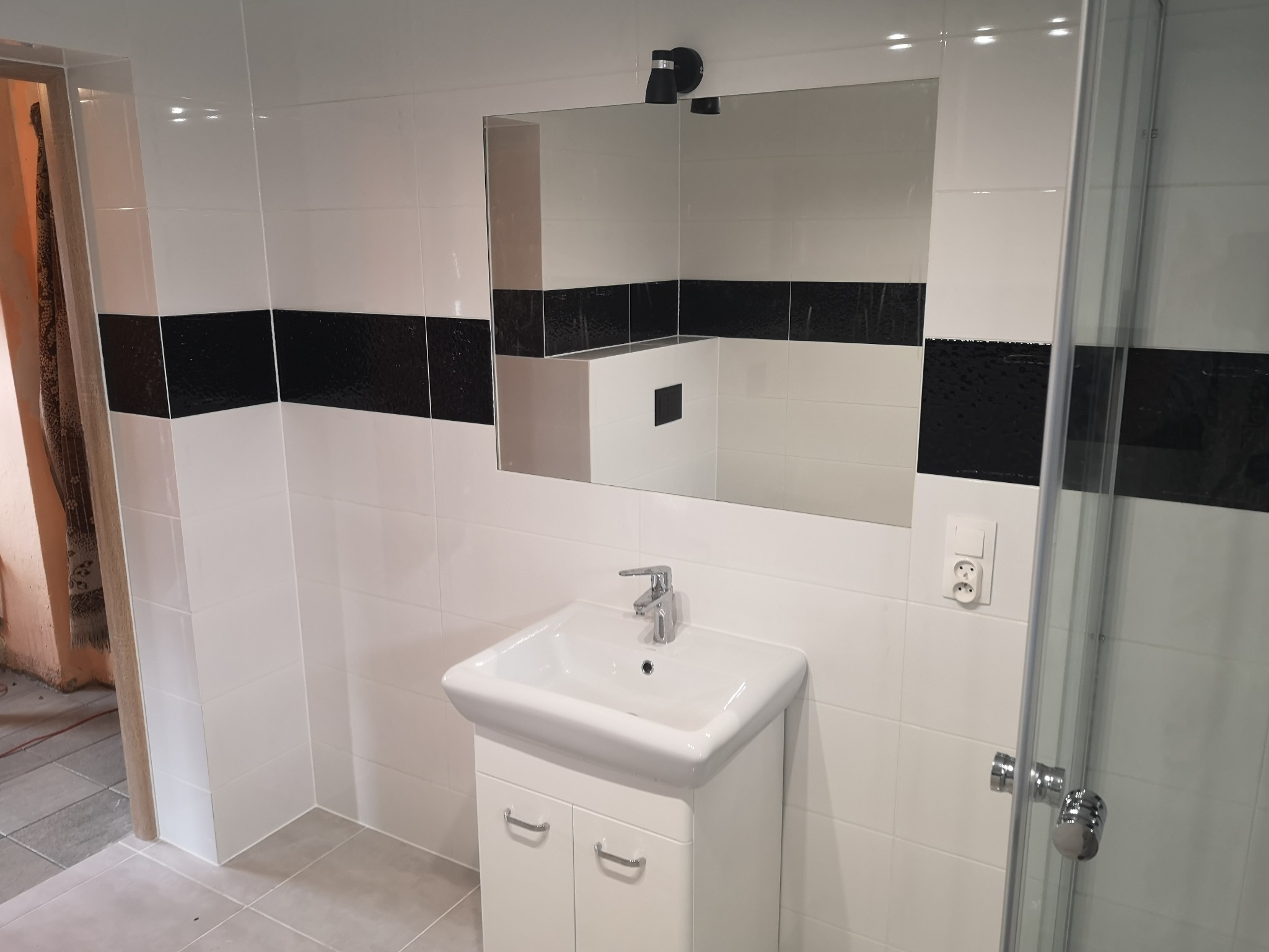 Nowoczesna łazienka z białymi i czarnymi płytkami, umywalką z szafką i kabiną prysznicową, widoczne szare płytki podłogowe i czarne dodatki.