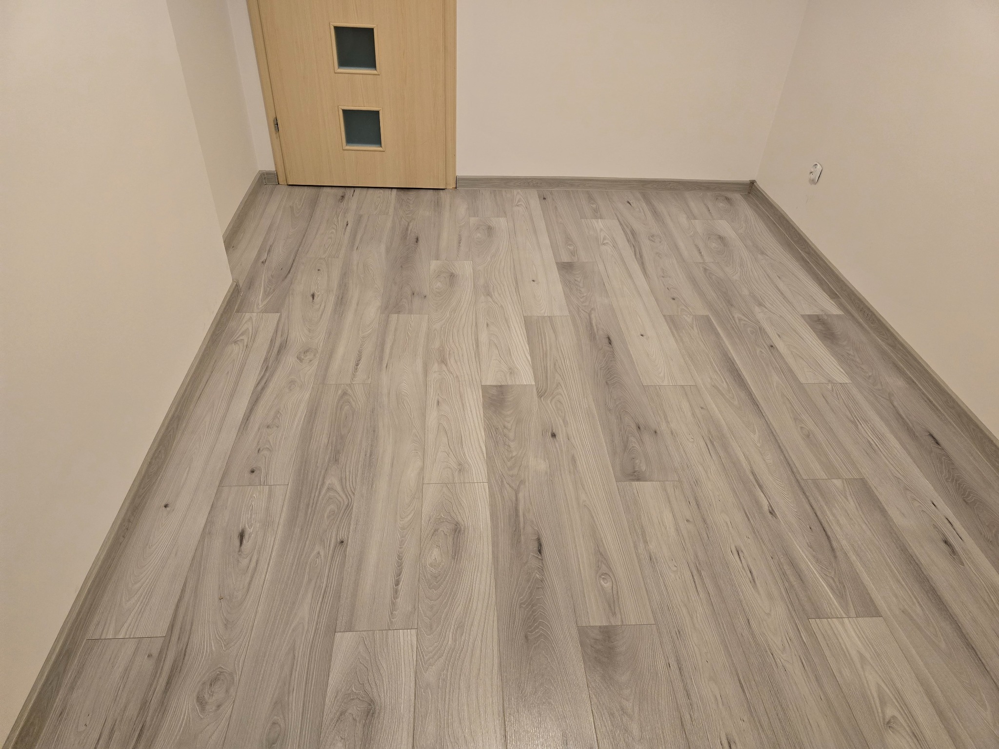 Jasnoszare panele podłogowe ułożone w nowym pomieszczeniu z drewnianymi drzwiami z dwoma kwadratowymi przeszkleniami i białą listwą przypodłogową.