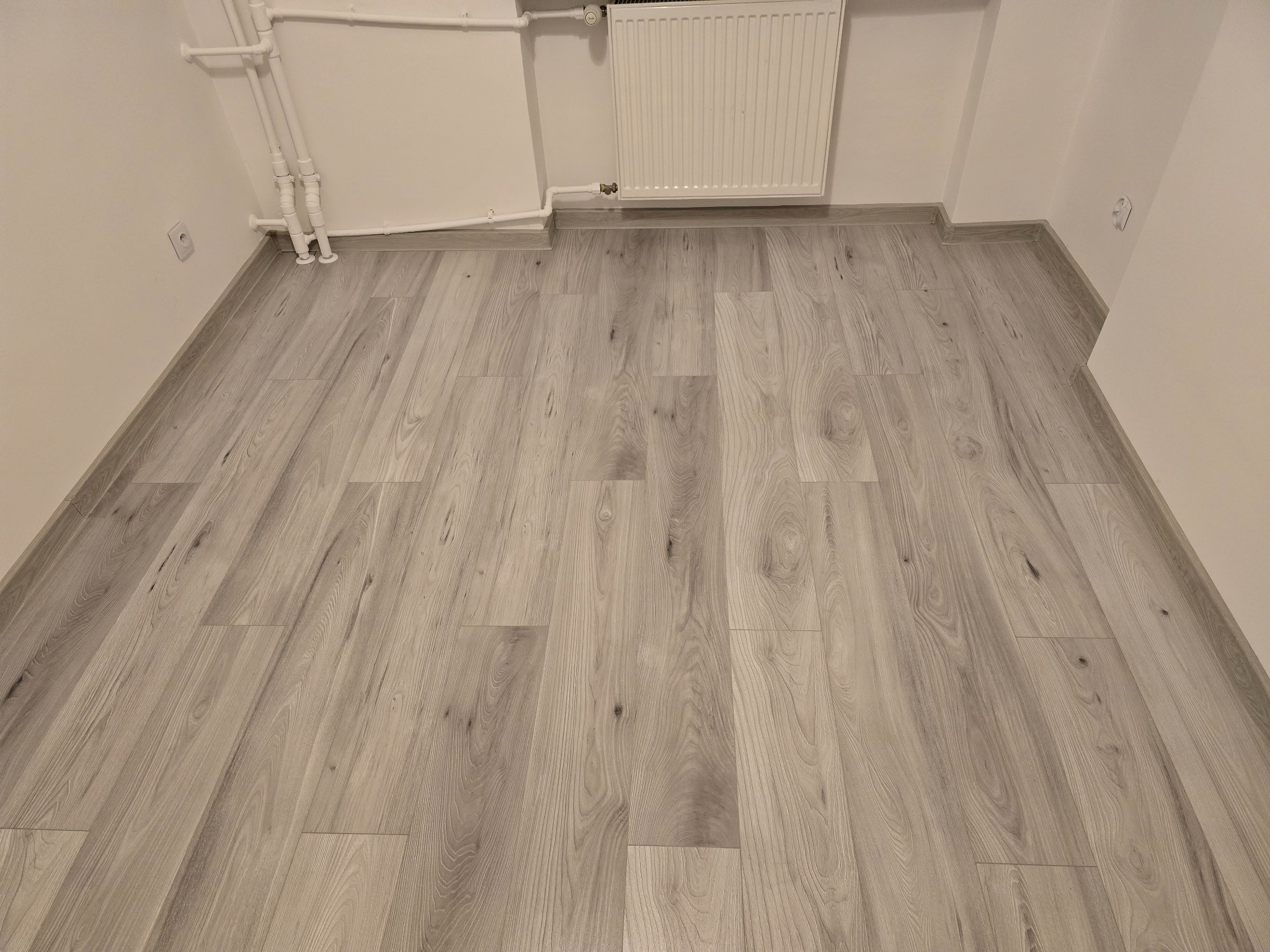 Jasnoszare panele podłogowe ułożone w nowym pomieszczeniu, widoczne listwy przypodłogowe, grzejnik i instalacja grzewcza na ścianie.