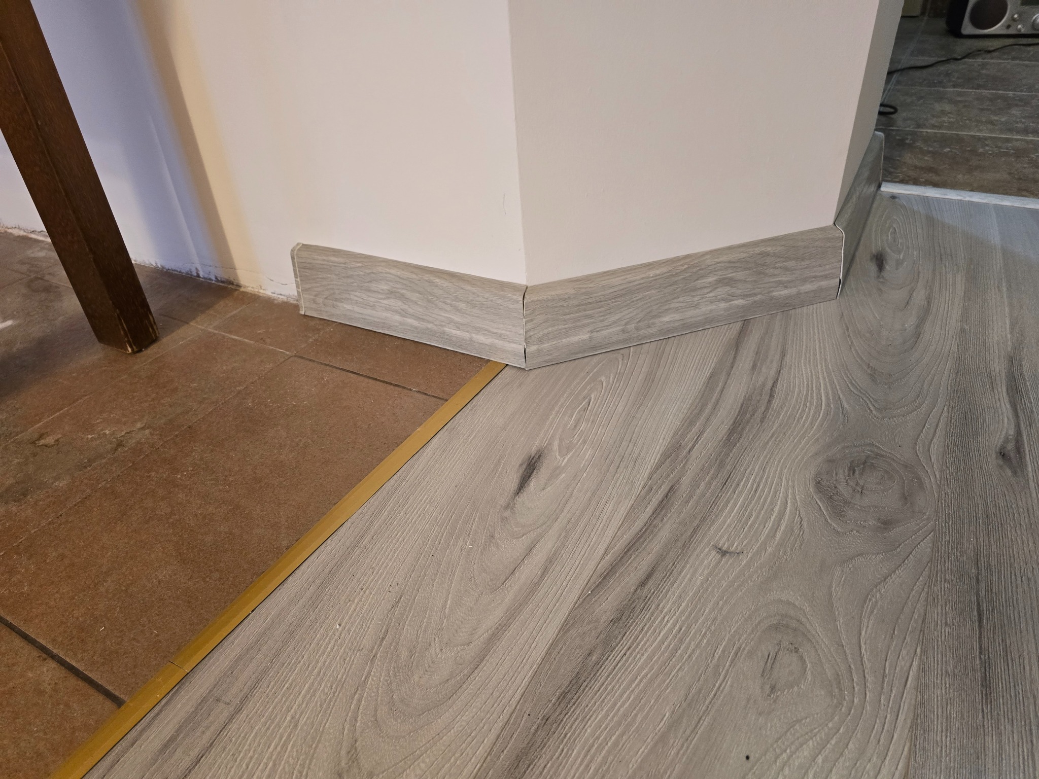Szczegółowe ujęcie połączenia paneli podłogowych imitujących drewno z płytkami ceramicznymi, listwa przypodłogowa w kolorze paneli, widoczny fragment nogi mebla.