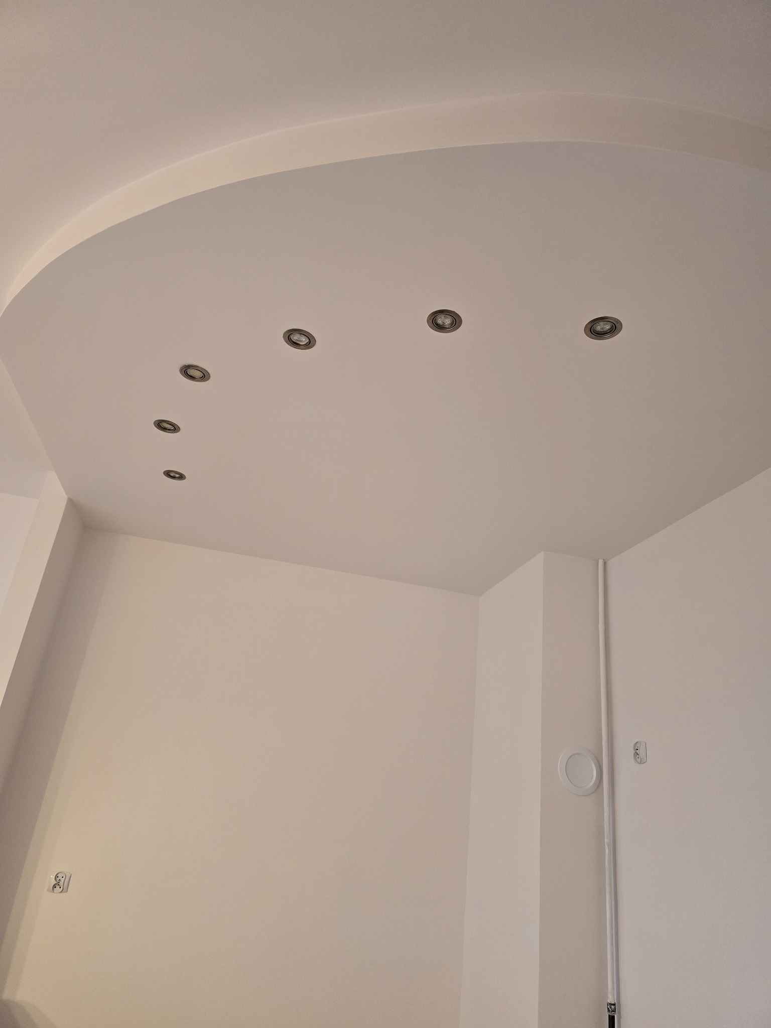 Biały sufit podwieszany z półokrągłym wycięciem i rzędem punktowych lamp sufitowych, widok z dołu na ścianę z gniazdkiem elektrycznym i pionową rurą.