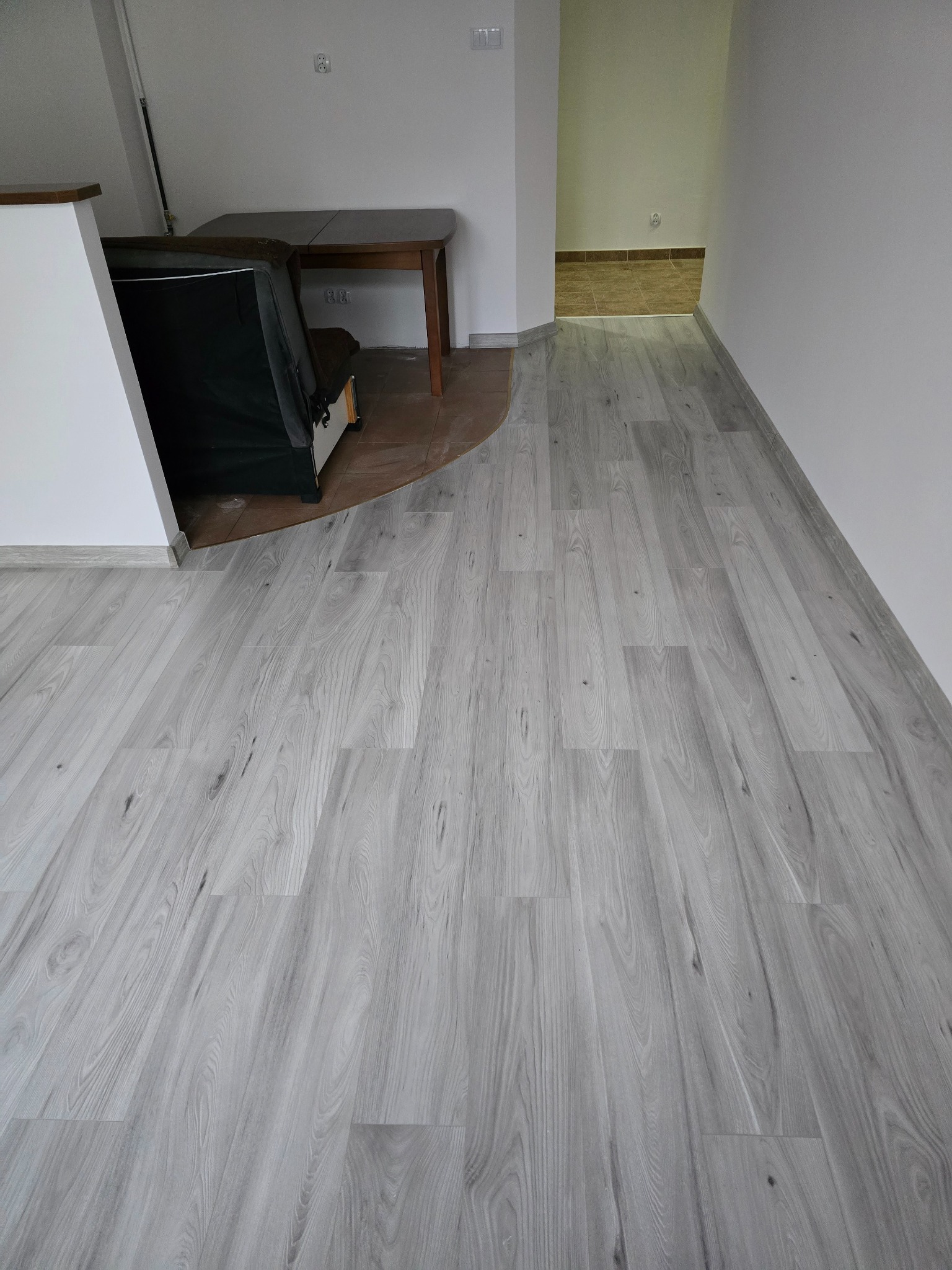 Jasnoszare panele podłogowe ułożone w nowym mieszkaniu, widoczny fragment sofy i stołu w tle.