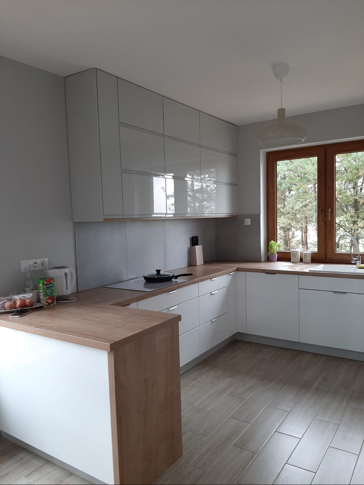 Jasna kuchnia z białymi, błyszczącymi szafkami górnymi i dolnymi, drewnianym blatem, widok na las za oknem.