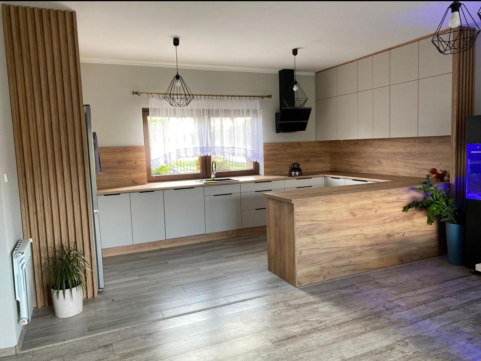 Nowoczesna, jasna kuchnia z drewnianymi akcentami: fronty szafek w kolorze złamanej bieli, drewniany panel ścienny, wyspa kuchenna z blatem imitującym drewno, wiszące lampy w stylu industrialnym...