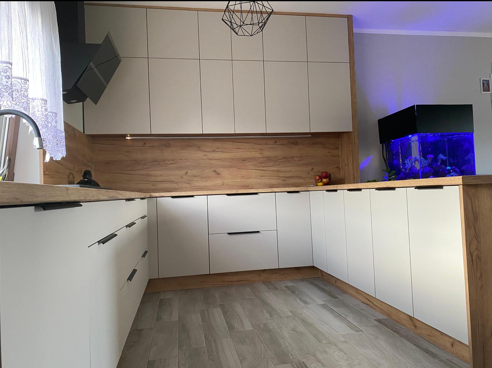 Nowoczesna kuchnia z białymi frontami i drewnianym blatem, oświetlona lampą wiszącą i podświetlonym akwarium, drewnopodobna okładzina ściany nad blatem.