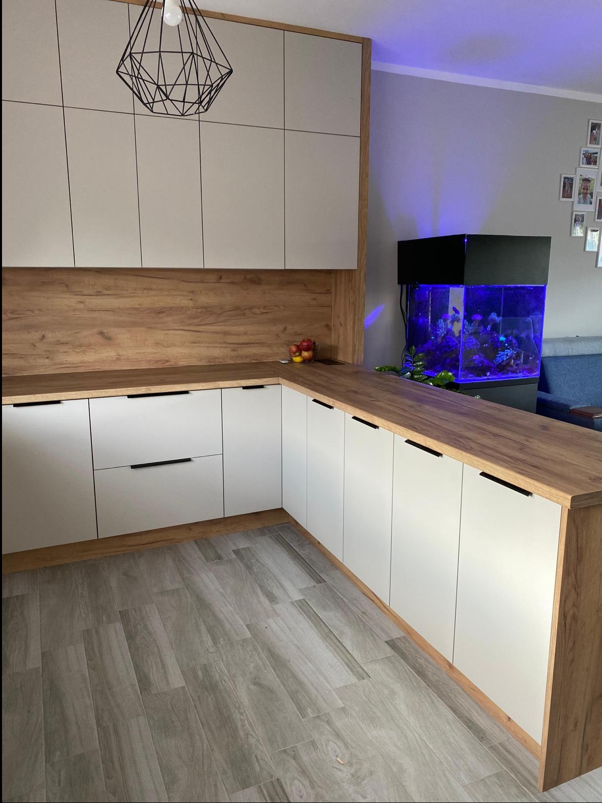 Nowoczesna kuchnia na wymiar z białymi frontami, drewnianym blatem i akcentami oraz akwarium z niebieskim oświetleniem w tle.