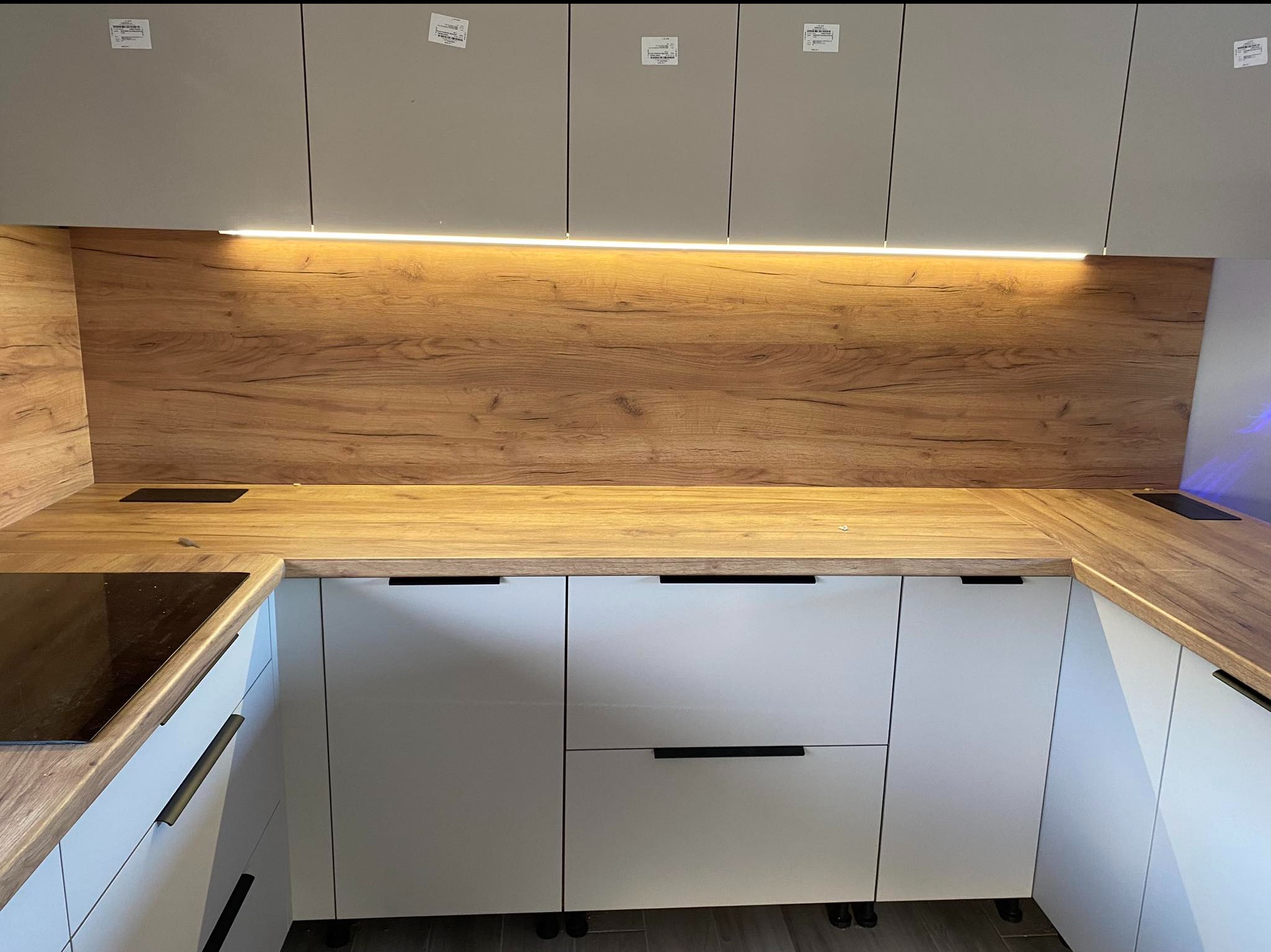 Nowoczesna kuchnia z białymi szafkami i drewnianym blatem, oświetlenie LED pod szafkami wiszącymi, płyta indukcyjna wkomponowana w blat.