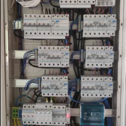 Otwarta skrzynka rozdzielcza z zamontowanymi bezpiecznikami, licznikiem energii elektrycznej i zabezpieczeniem przeciwprzepięciowym, widoczne okablowanie w kolorach niebieskim, czarnym...