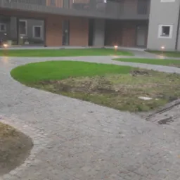 Oświetlenie ścieżki wzdłuż budynku mieszkalnego, lampy ogrodowe włączone, kostka brukowa, trawnik.