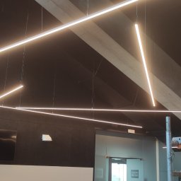 EL-HOUSE DAMIAN NOWAKOWSKI - Nowoczesne oświetlenie biurowe: geometryczny układ liniowych lamp LED zawieszonych na tle ciemnej ściany i sufitu, z widocznym rusztowaniem w tle, sugerującym trwający montaż.