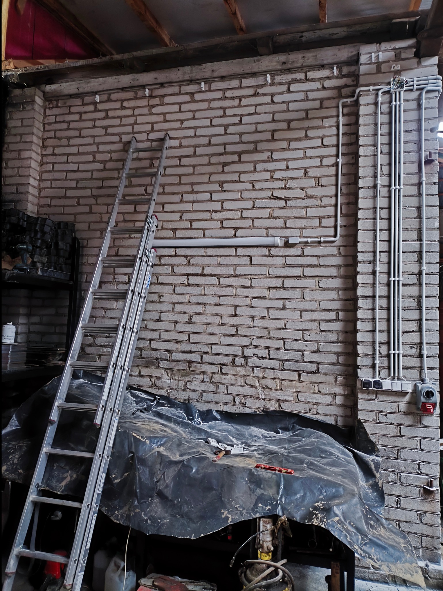 Murowana ściana z widoczną instalacją elektryczną w rurkach, drabina oparta o ścianę, zabezpieczona folią podłoga, narzędzia na folii, remontowane pomieszczenie.
