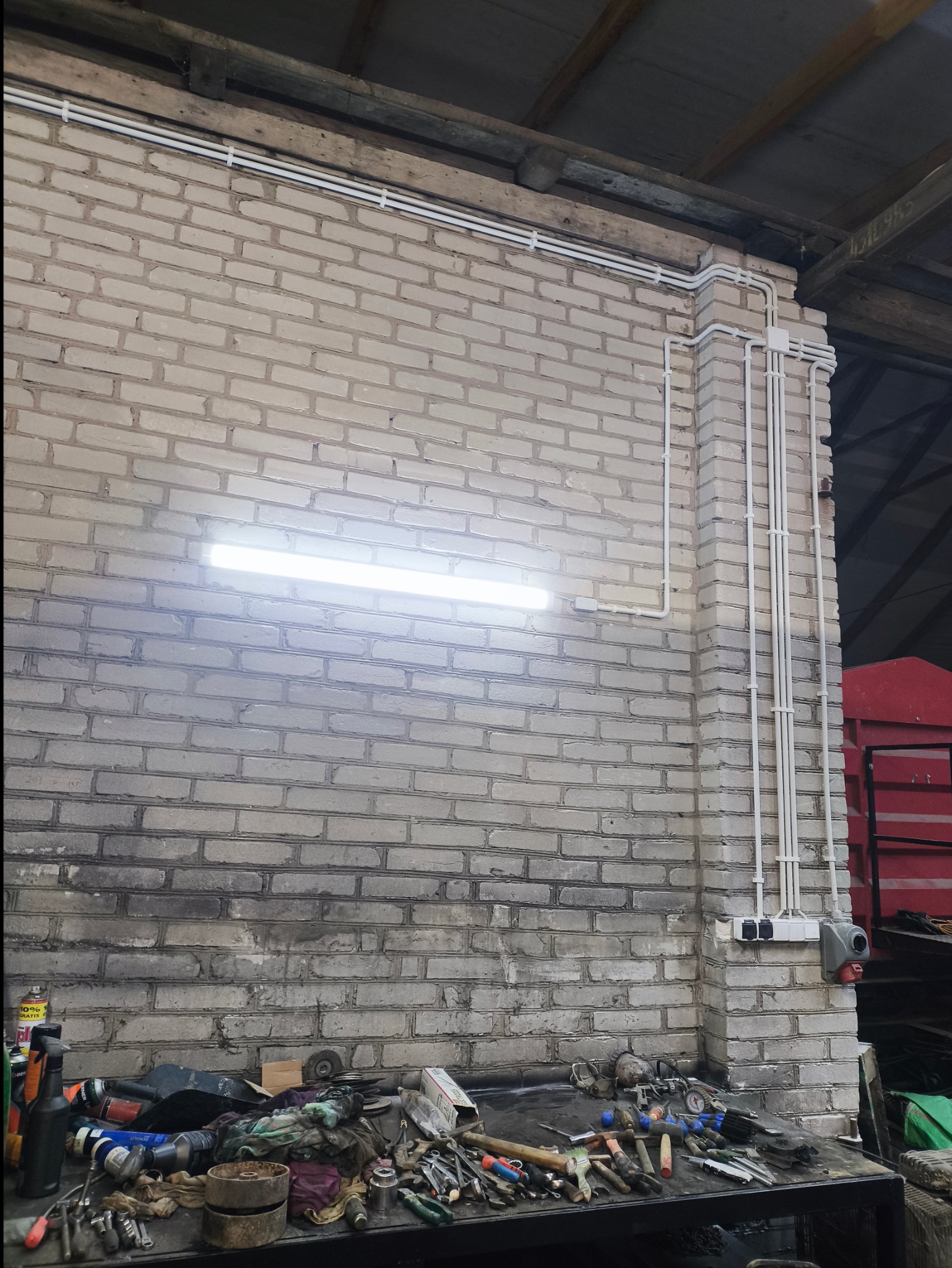 Biała ściana z cegły z widoczną instalacją elektryczną w postaci rurek PCV, oświetleniem jarzeniowym i gniazdkami. Na pierwszym planie stół warsztatowy z narzędziami i akcesoriami.