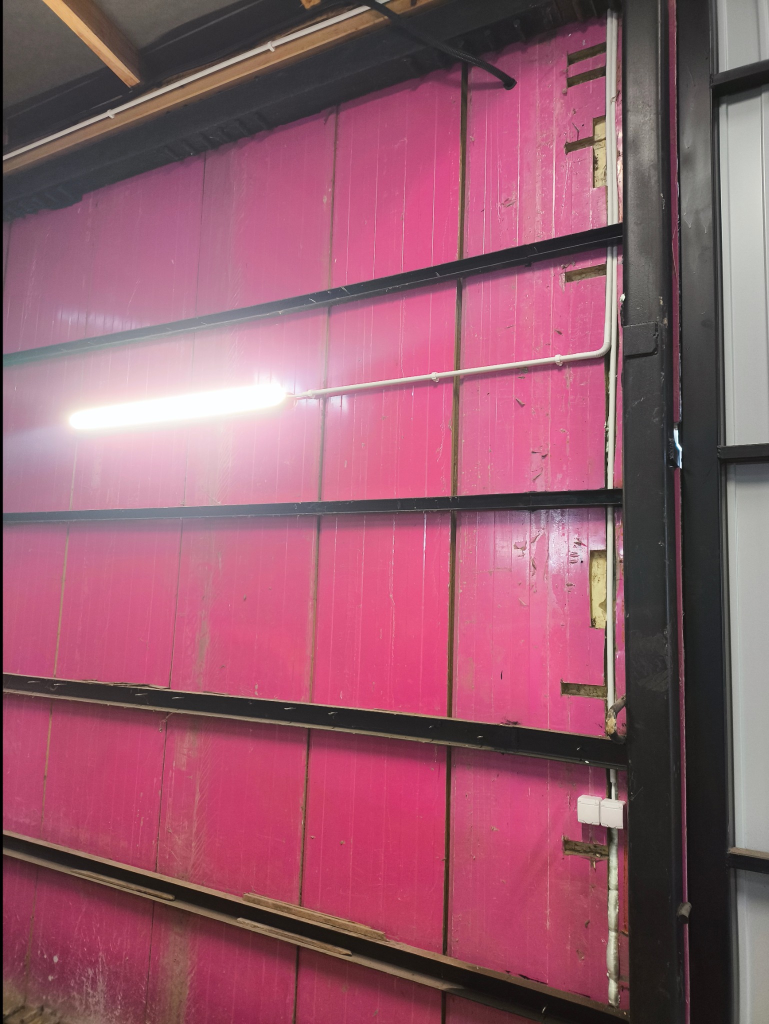 Wnętrze hali produkcyjnej z różową ścianą z płyt warstwowych, widoczne metalowe profile konstrukcyjne, białe rury instalacji elektrycznej i pojedyncza świetlówka.