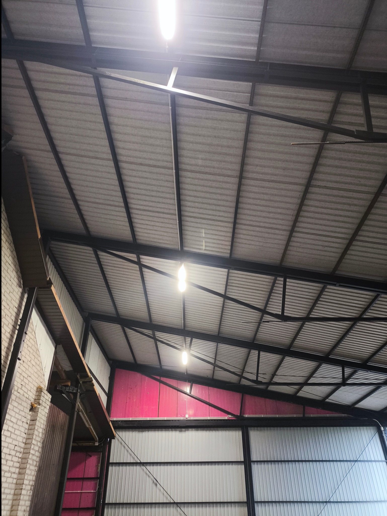 Wnętrze hali z widoczną konstrukcją stalową dachu, oświetleniem LED i częściowo wykończonymi ścianami w kolorze szarym i różowym.