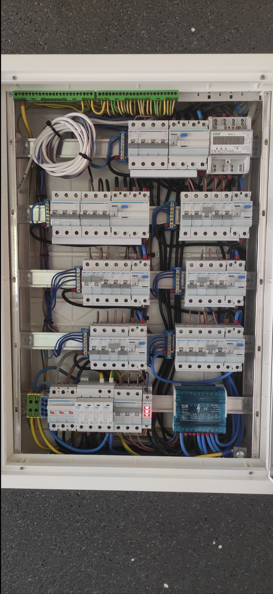 Otwarta skrzynka rozdzielcza z zamontowanymi bezpiecznikami, licznikiem energii elektrycznej i zabezpieczeniem przeciwprzepięciowym, widoczne okablowanie w kolorach niebieskim, czarnym...