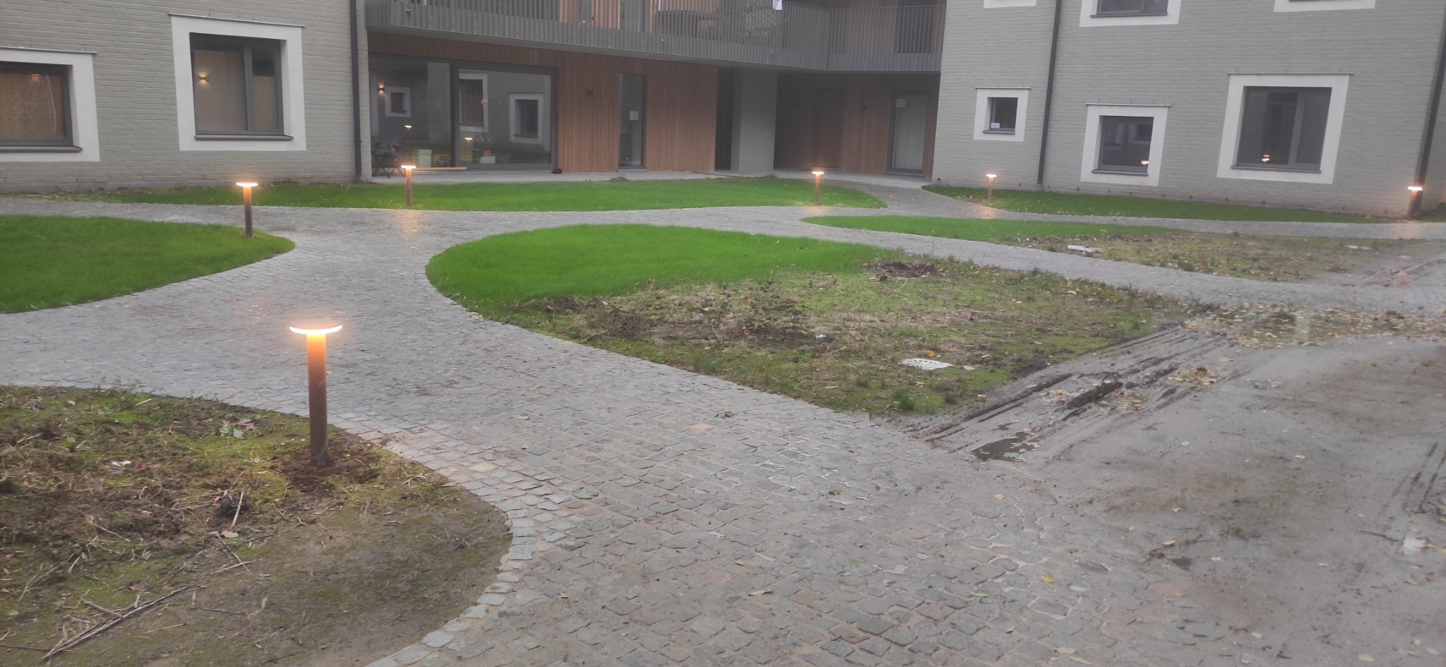 Oświetlenie ścieżki wzdłuż budynku mieszkalnego, lampy ogrodowe włączone, kostka brukowa, trawnik.