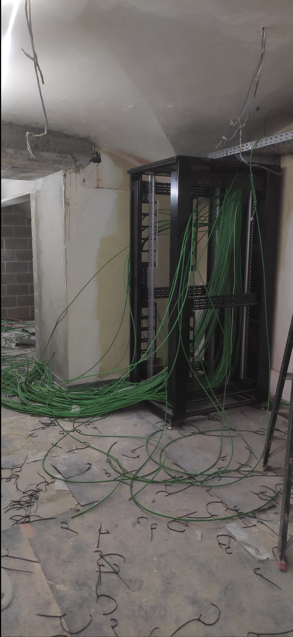 Niedokończona instalacja serwerowni: czarna szafa rack z zielonymi kablami w surowym pomieszczeniu z widocznymi przewodami pod sufitem i metalowymi elementami na podłodze.