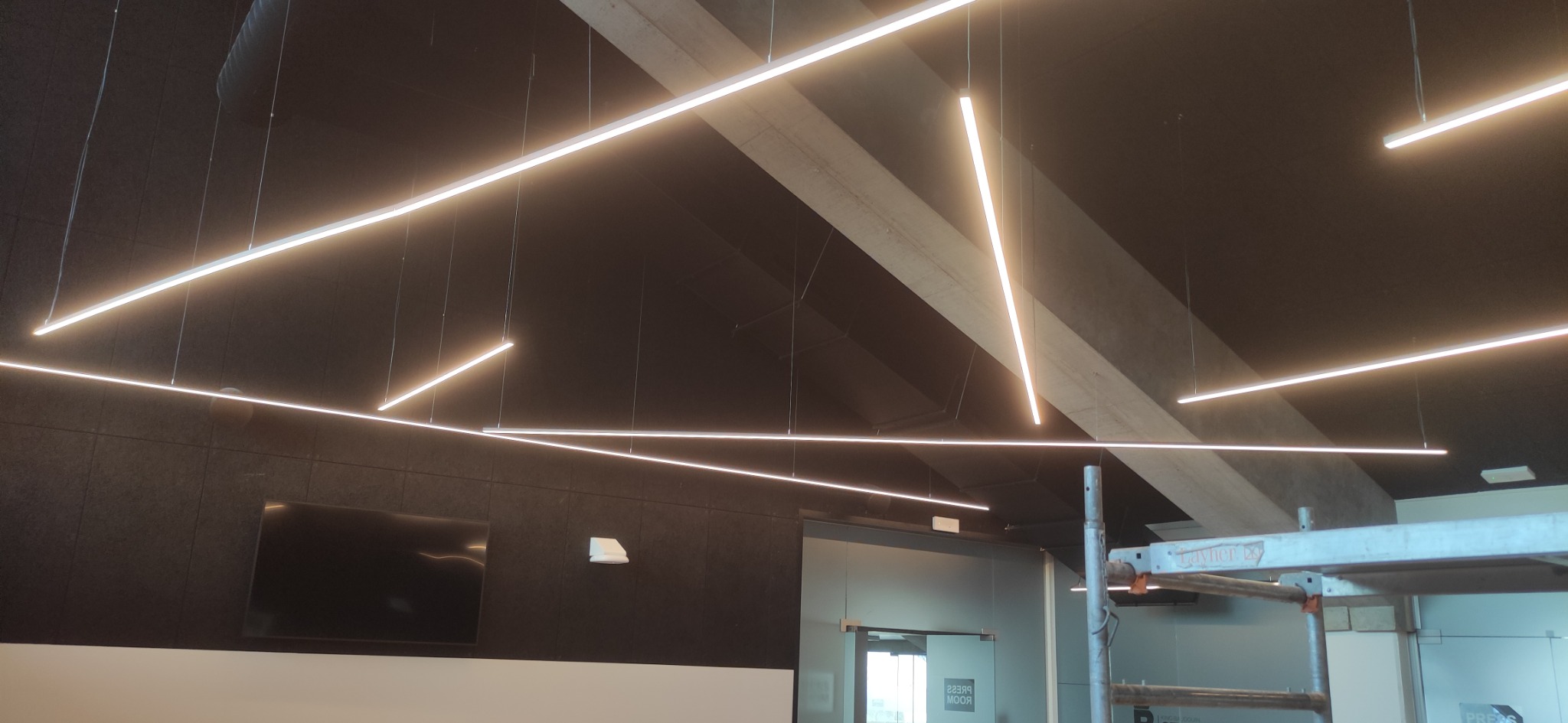 Nowoczesne oświetlenie biurowe: geometryczny układ liniowych lamp LED zawieszonych na tle ciemnej ściany i sufitu, z widocznym rusztowaniem w tle, sugerującym trwający montaż.