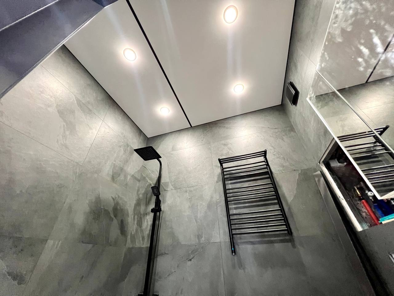 Nowoczesna łazienka z szarymi płytkami imitującymi marmur, czarną armaturą, sufit podwieszany z wpuszczonymi lampami i grzejnikiem drabinkowym.