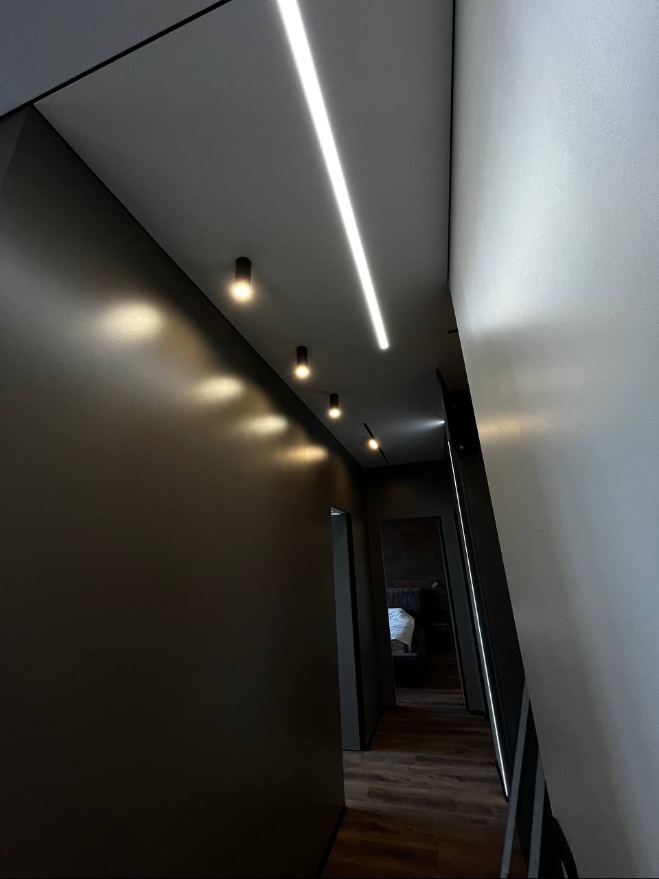 Nowoczesny korytarz z ciemnymi ścianami, oświetleniem punktowym i liniowym LED na suficie, widok w głąb mieszkania z drewnianą podłogą.