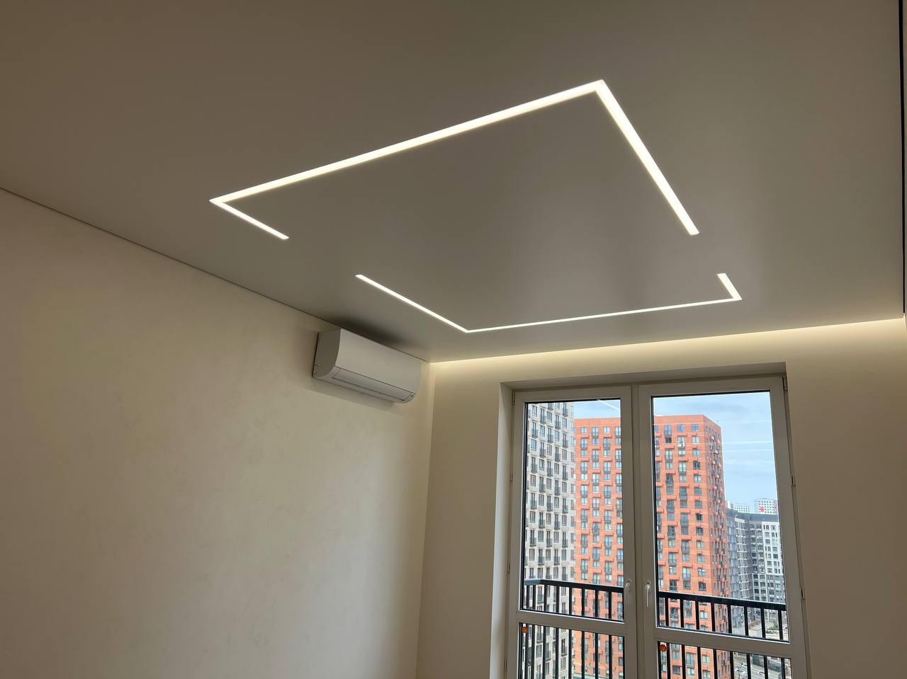 Pokój z białym sufitem napinanym i wbudowanym oświetleniem LED w kształcie kwadratu i linii, klimatyzator na ścianie, widok z okna na budynki.