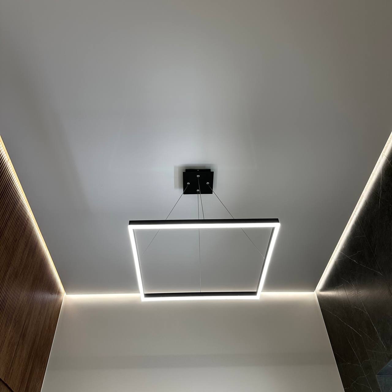 Nowoczesny salon z podwieszanym sufitem, oświetleniem LED wzdłuż krawędzi i kwadratową lampą wiszącą z białym światłem.