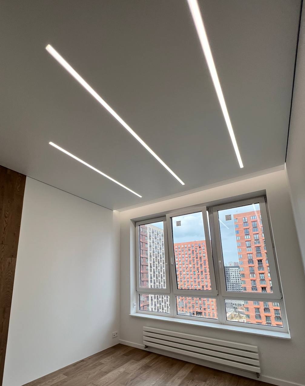Pokój z trzema liniowymi lampami LED w suficie napinanym, widok na budynki mieszkalne za oknem.