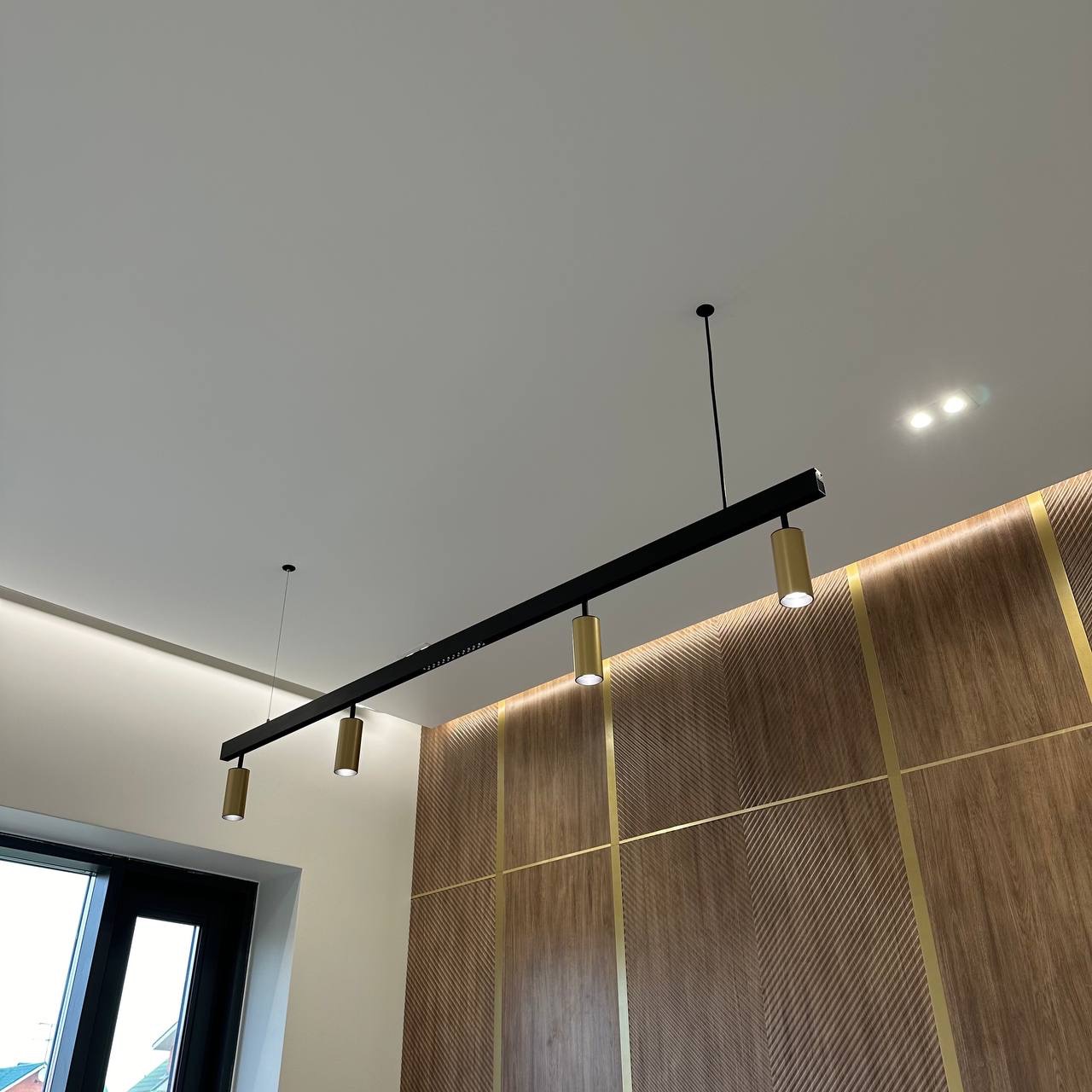 Nowoczesne oświetlenie sufitowe: czarna szyna z złotymi reflektorami, drewniana ściana z geometrycznym wzorem i złotymi akcentami.